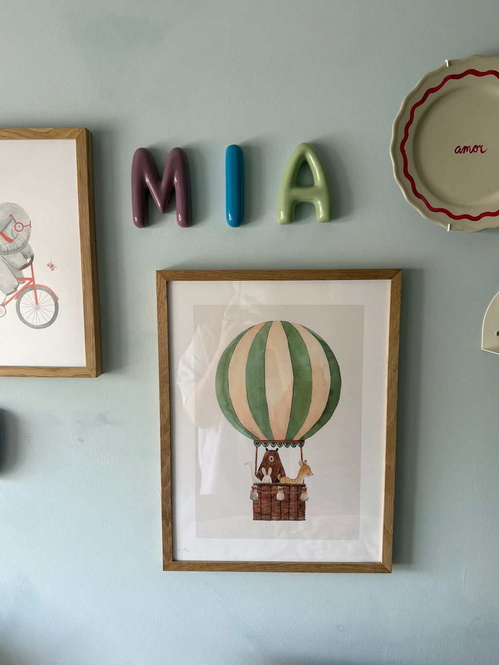 A styled interior wall with framed art and decorative objects. The central framed piece shows a vintage-style hot air balloon illustration in green, cream, and brown, mounted on a pale blue wall.