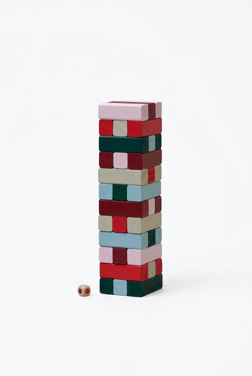 A vertical stack of colorful wooden blocks on a plain white background, with a small die placed at the base. The blocks come in multiple colors including red, green, beige, light blue, and pink.