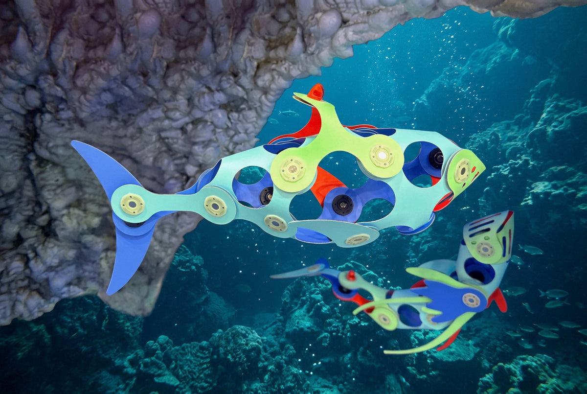 Colorful robotic fish toys swimming in an underwater coral reef scene. The main fish toy is light blue with circular cutouts showing internal gears and colorful accents, while a smaller matching fish toy swims below it.