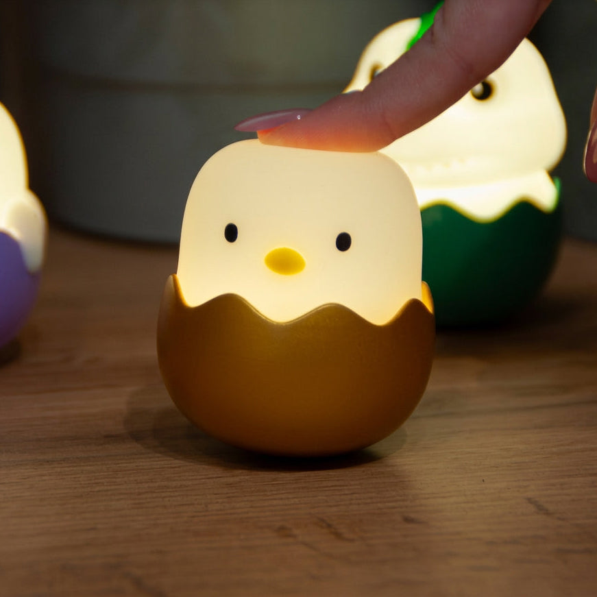 A cute egg-shaped night light shaped like a chick hatching from an egg, glowing with a warm yellow light. A human hand is gently touching the top of the chick's head, and another similar night light is blurred in the background on a wooden surface.