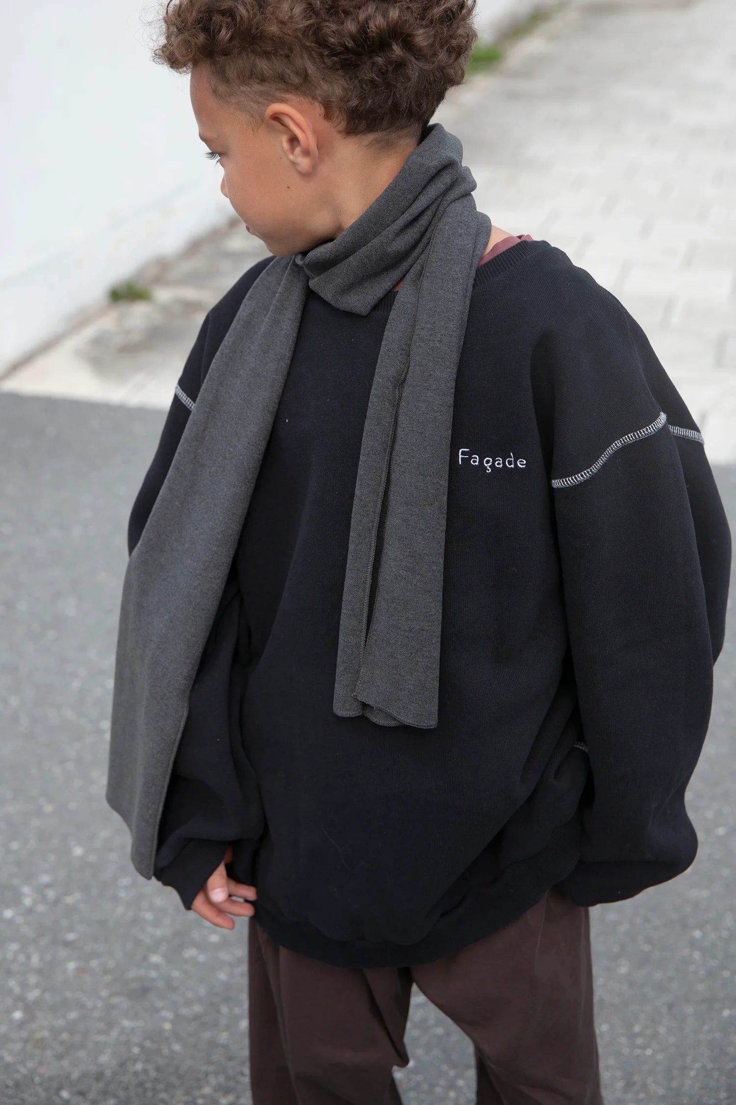 A child wearing a black oversized sweatshirt with a gray knit scarf, brown pants, and white contrast stitching details, standing outdoors.