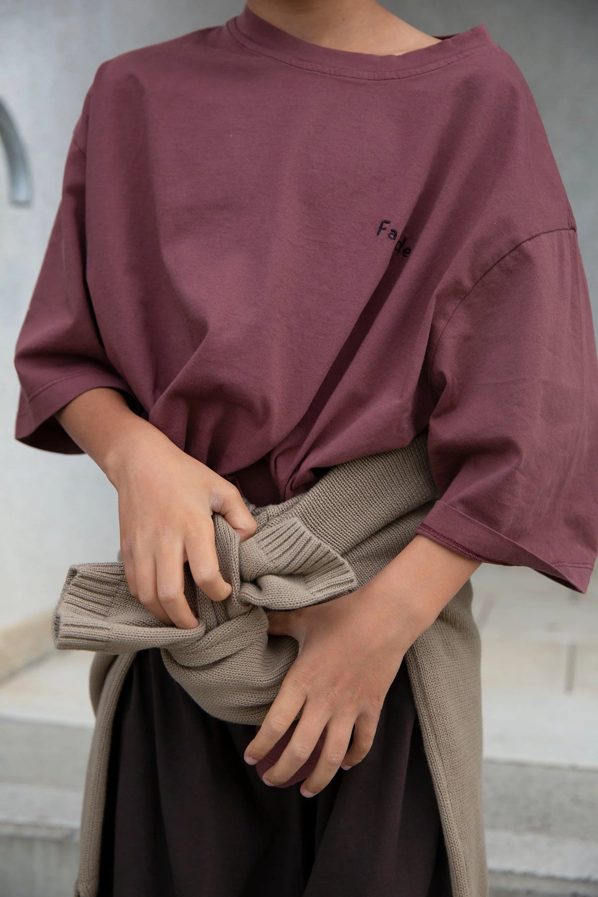 A person wearing an oversized dusty mauve t-shirt with a small black embroidered logo on the chest, holding a taupe knit sweater tied around their waist.
