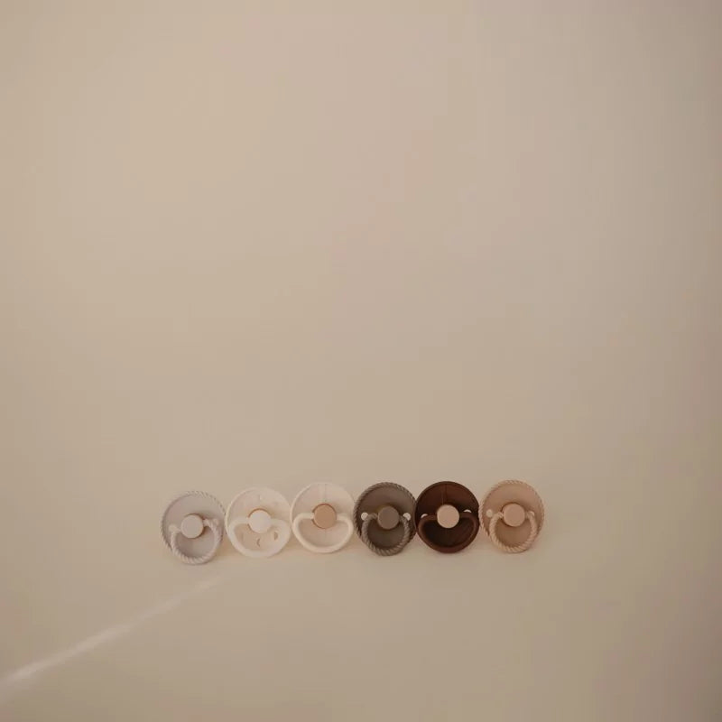 Set of five baby pacifiers in different colors arranged in a row on a beige background.