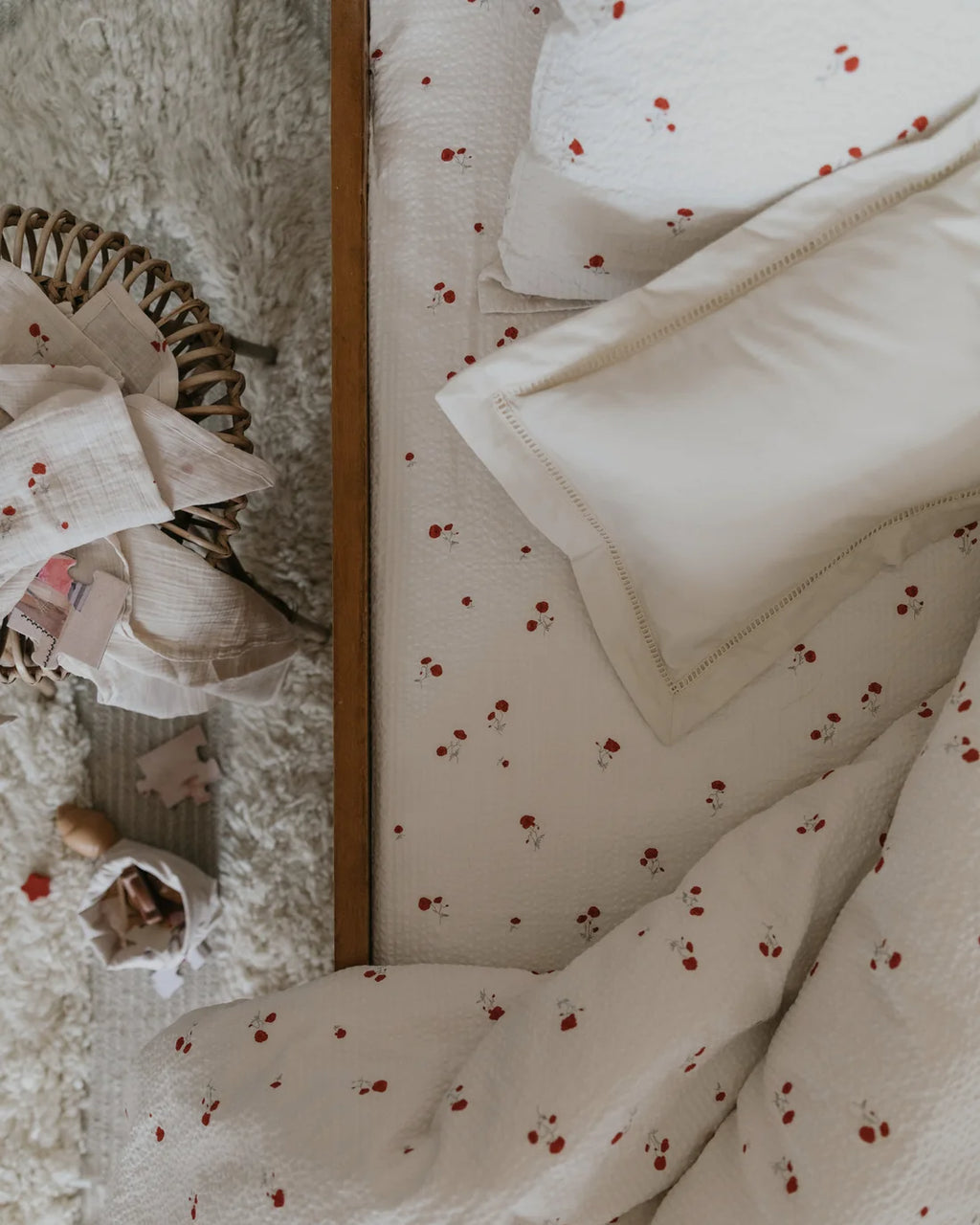 A cozy bedroom scene showing a bed dressed in cream bedding with a small red cherry print, alongside matching pillows and a wooden headboard. A mirror on the left reflects a soft, styled room corner with folded linens and toys.