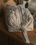 A quilted cream-colored cosmetic pouch with a small floral print, resting on a wooden surface in warm, natural light.
