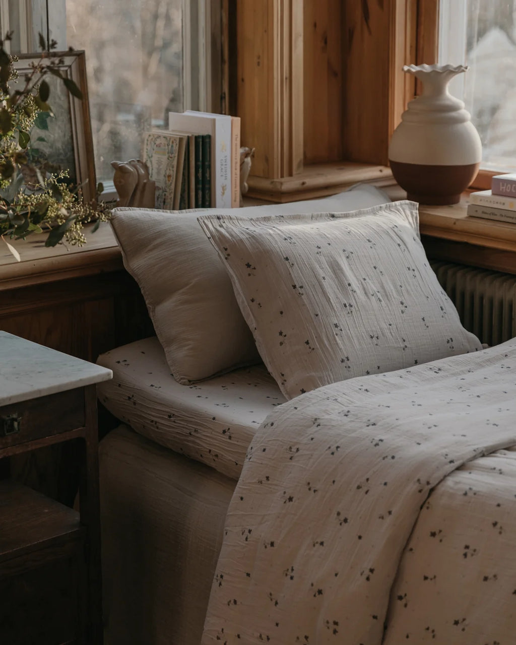 A neatly made bed in a rustic bedroom setting, featuring a light beige cotton muslin bedding set with a delicate dark floral print. The bed is styled with matching pillows and a duvet cover, set against a warm wood-paneled background with a window, books, and a ceramic vase.