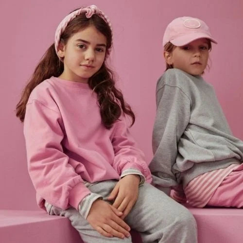 Two children wearing matching pink and gray casual outfits, seated on pink blocks against a solid pink studio background.