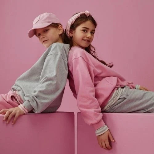 Two children sitting back-to-back on pink blocks against a pink background. One child wears a light gray sweatshirt, pink pants, and a pink cap, while the other wears a pink sweatshirt, gray pants, and a pink headband.