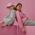 Two children sitting back-to-back on pink blocks against a pink background. One child wears a light gray sweatshirt, pink pants, and a pink cap, while the other wears a pink sweatshirt, gray pants, and a pink headband.