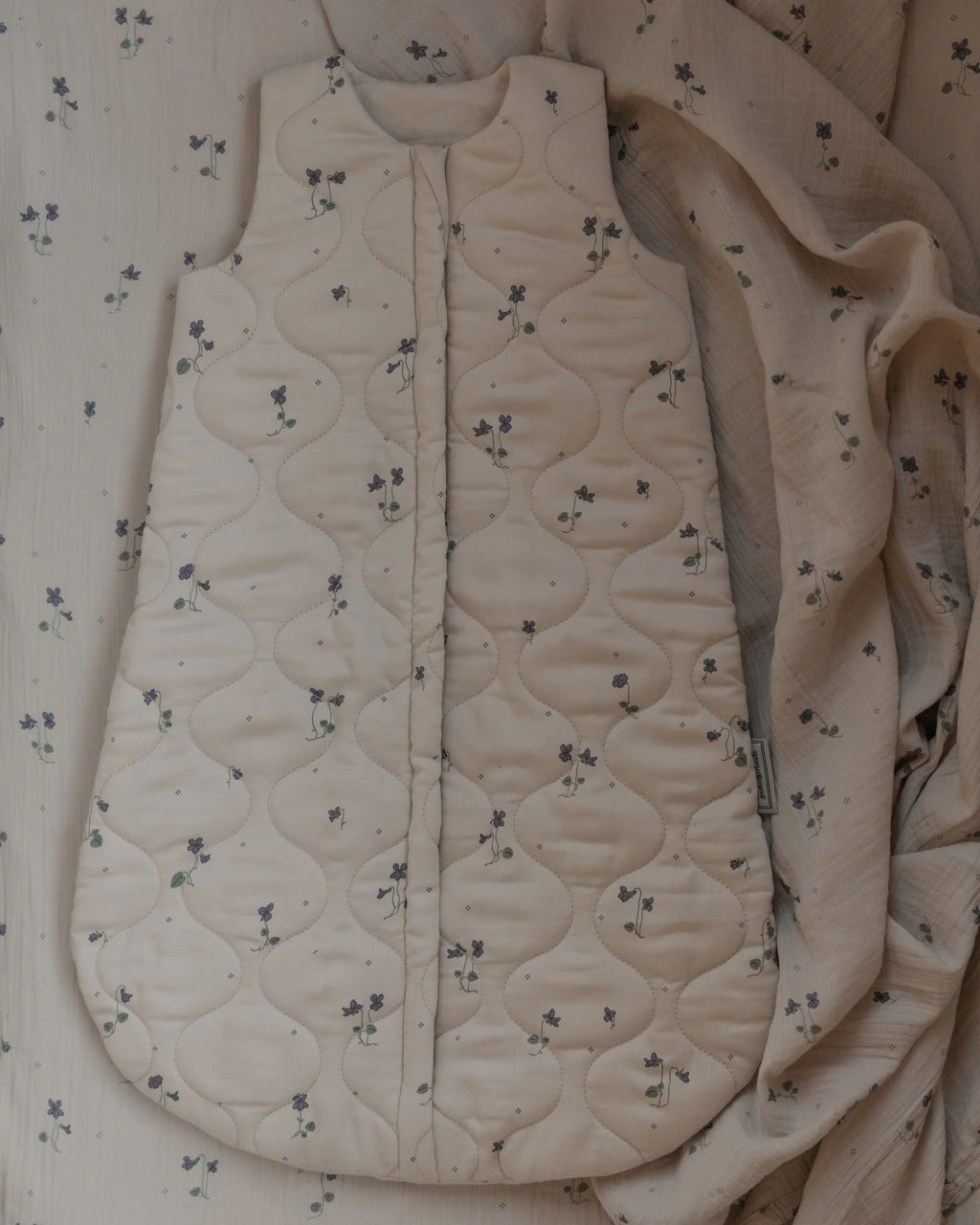 off-white quilted baby sleep sack with a small purple floral pattern, featuring a front zipper closure and a side tag.