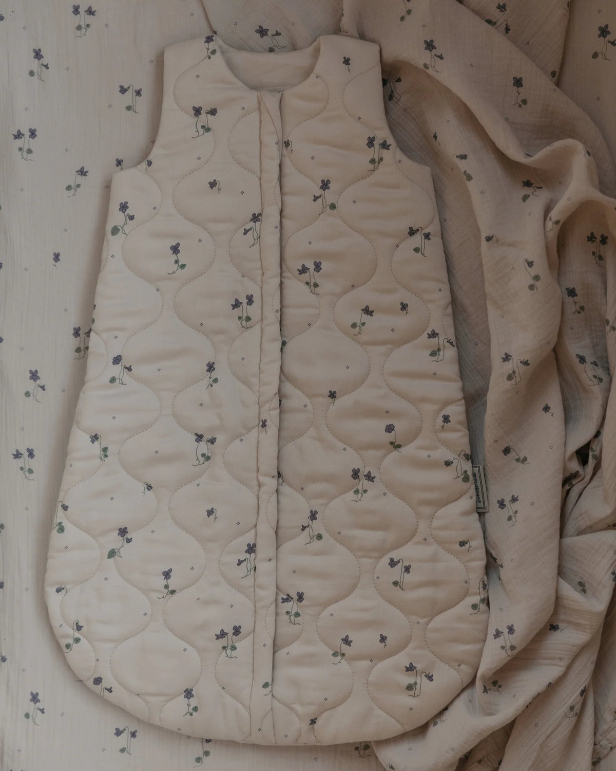 off-white quilted baby sleep sack with a small purple floral pattern, featuring a front zipper closure and a side tag.