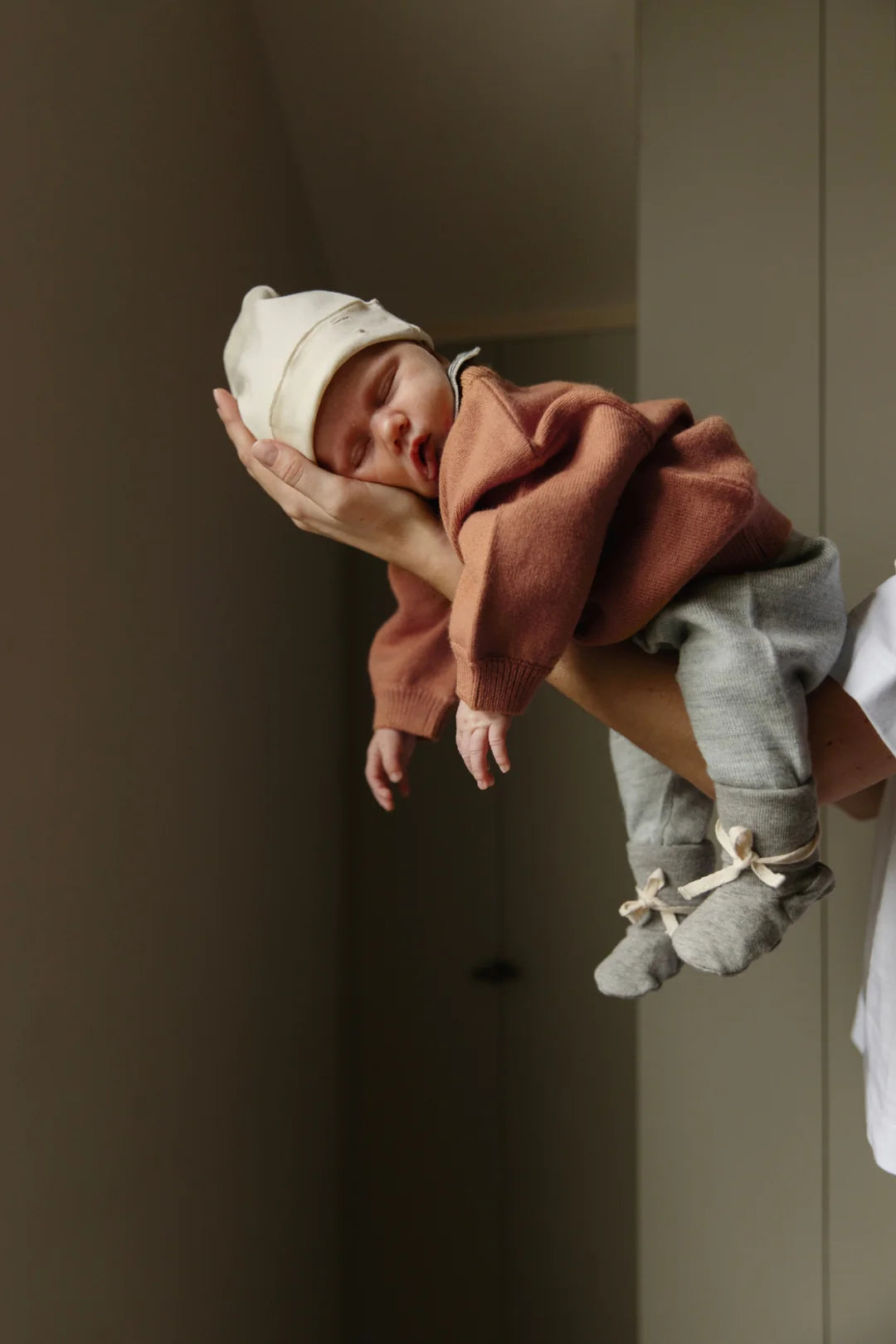 A newborn baby wearing a cream knit hat, a rust-brown knit sweater, light gray pants, and gray booties with ribbon bows, held gently in the air by an adult's hands against a soft beige background.