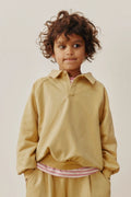A young child with curly brown hair wearing a mustard yellow long-sleeve collared sweatshirt and matching pants, standing against a plain light beige background.