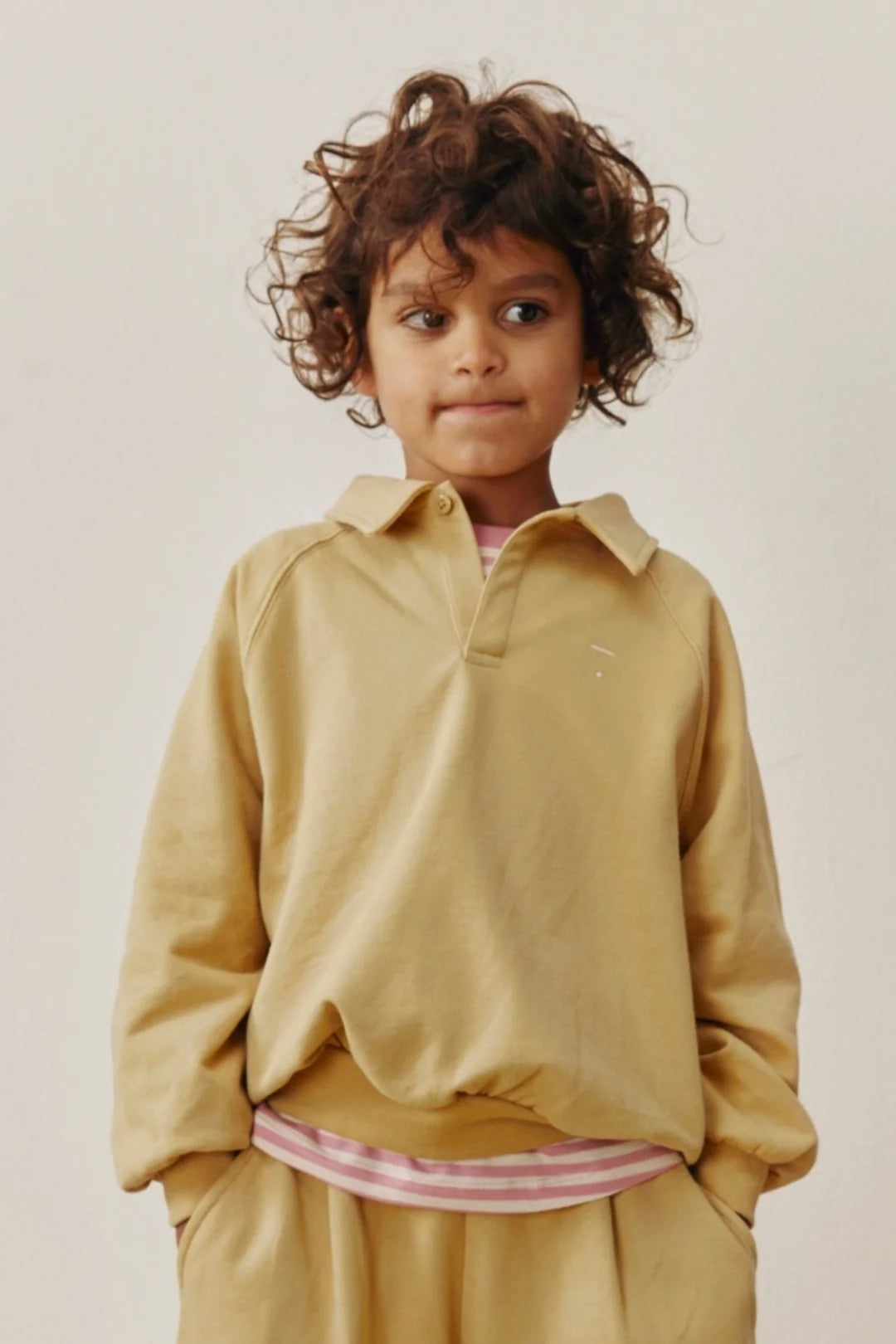 A young child with curly brown hair wearing a mustard yellow long-sleeve collared sweatshirt and matching pants, standing against a plain light beige background.