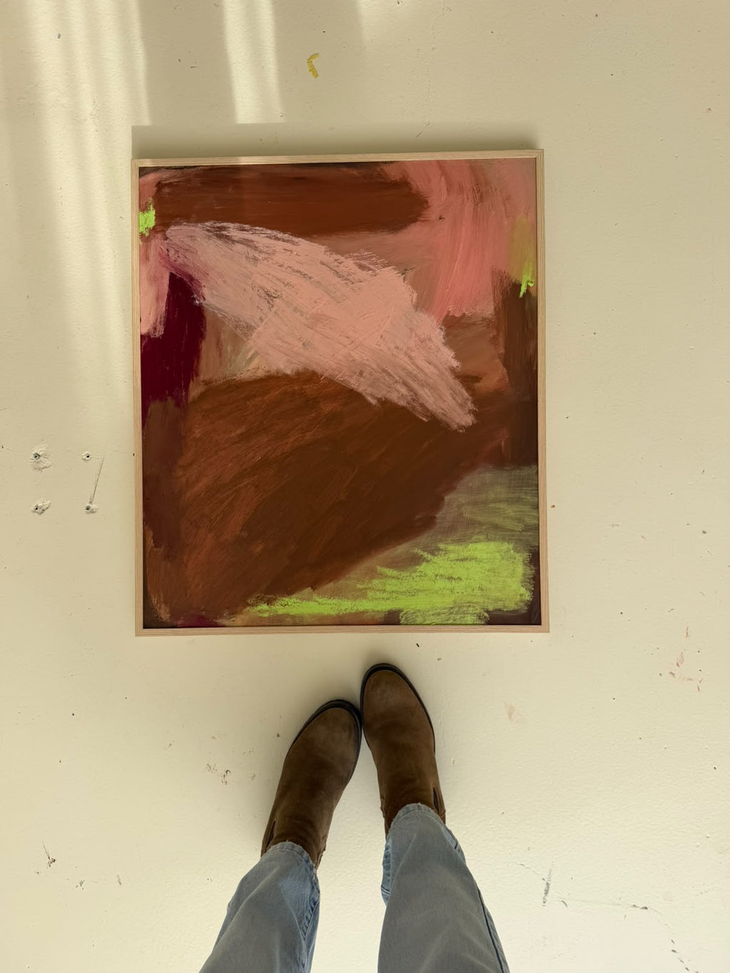 An abstract framed painting with pink, brown, burgundy, and lime green brushstrokes, photographed from above on a white floor with a person's legs and boots visible at the bottom.