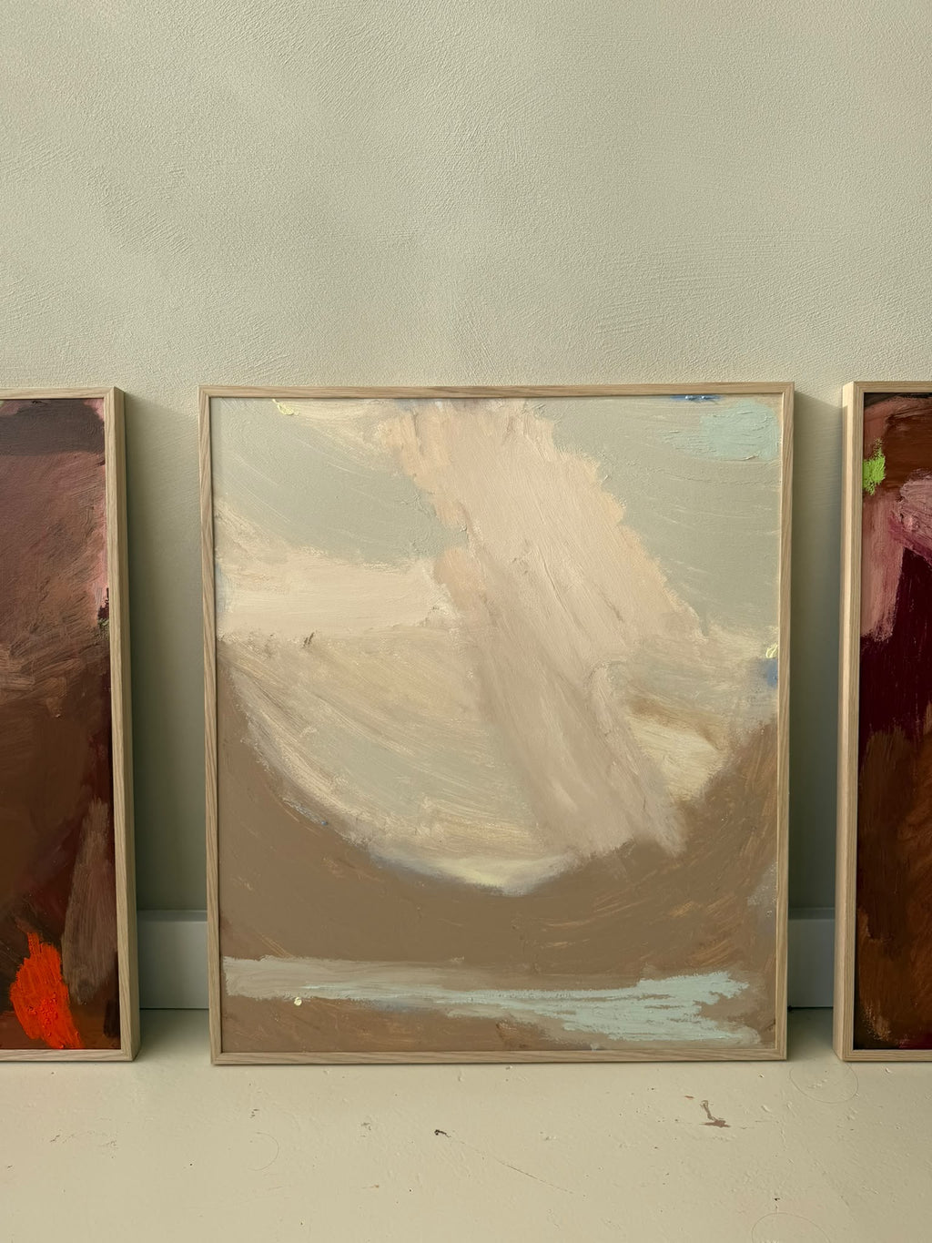 A framed abstract landscape painting with soft brushstrokes in beige, brown, cream, and light blue, leaning against a light gray wall. Partially visible paintings flank the central framed piece.