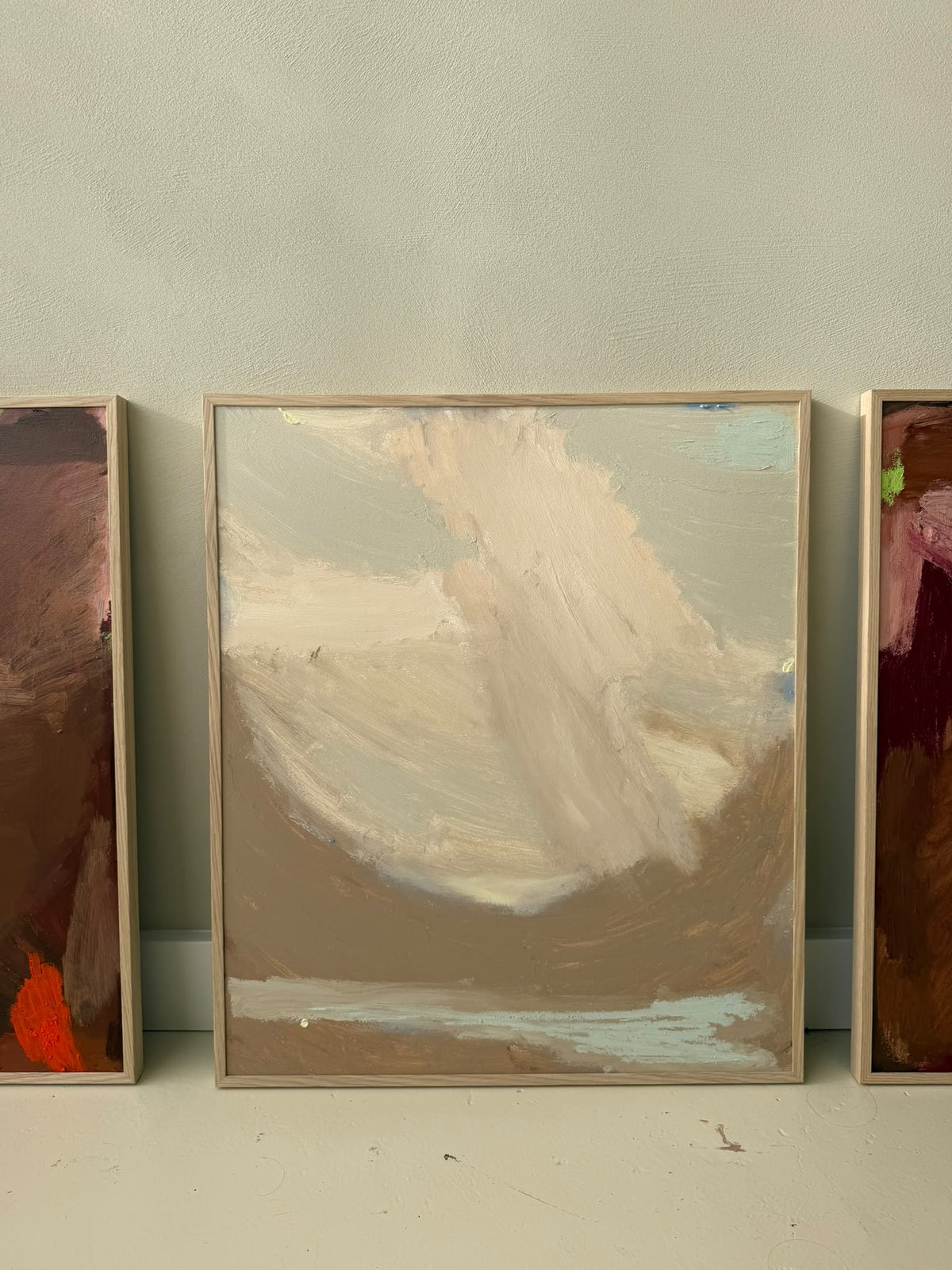 A framed abstract landscape painting with soft brushstrokes in beige, brown, cream, and light blue, leaning against a light gray wall. Partially visible paintings flank the central framed piece.