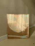 A framed abstract landscape painting leaning against a beige wall on a floor. The artwork features sweeping brushstrokes of cream, tan, and brown, with a light wood frame.