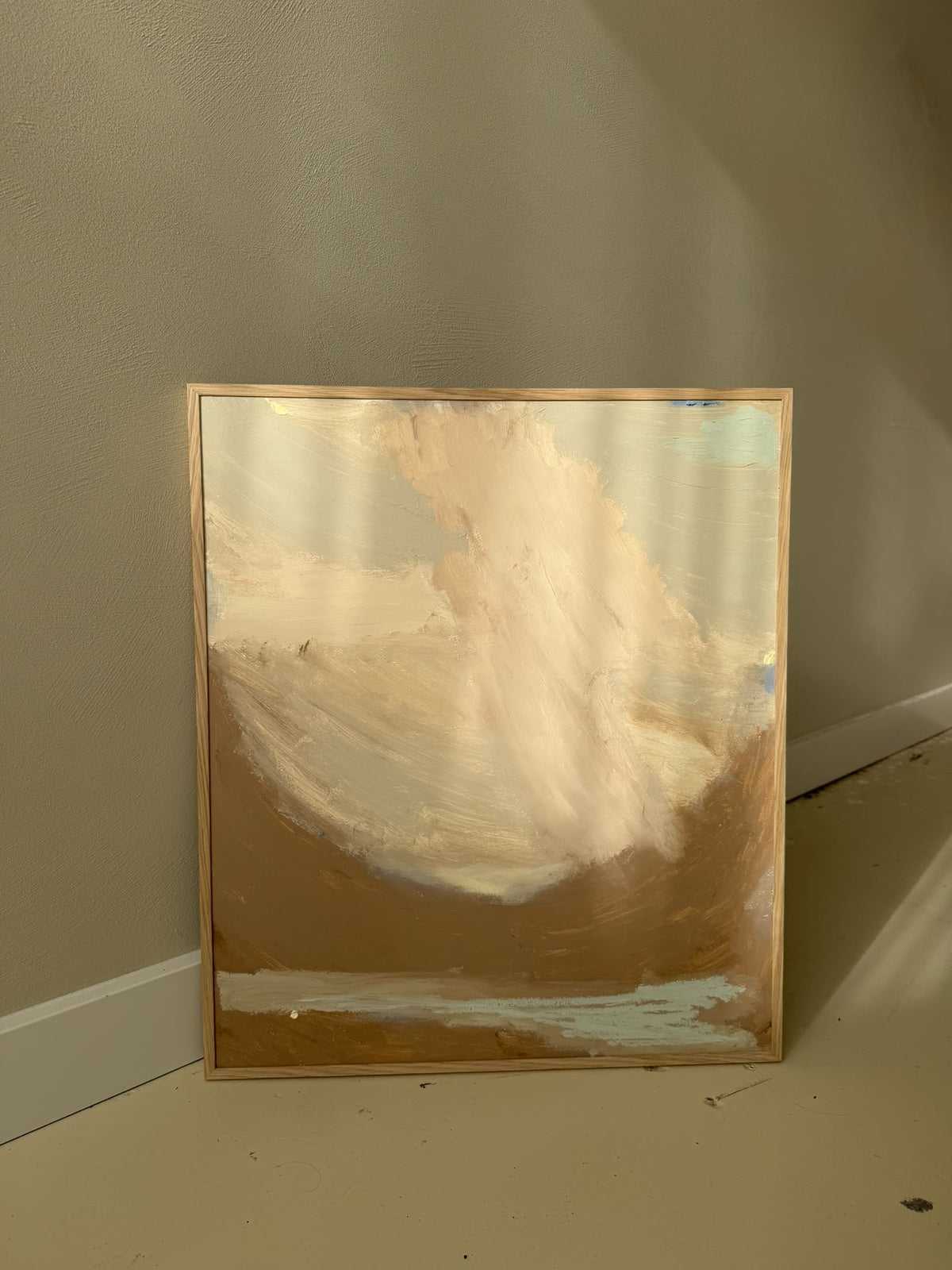 A framed abstract landscape painting leaning against a beige wall on a floor. The artwork features sweeping brushstrokes of cream, tan, and brown, with a light wood frame.