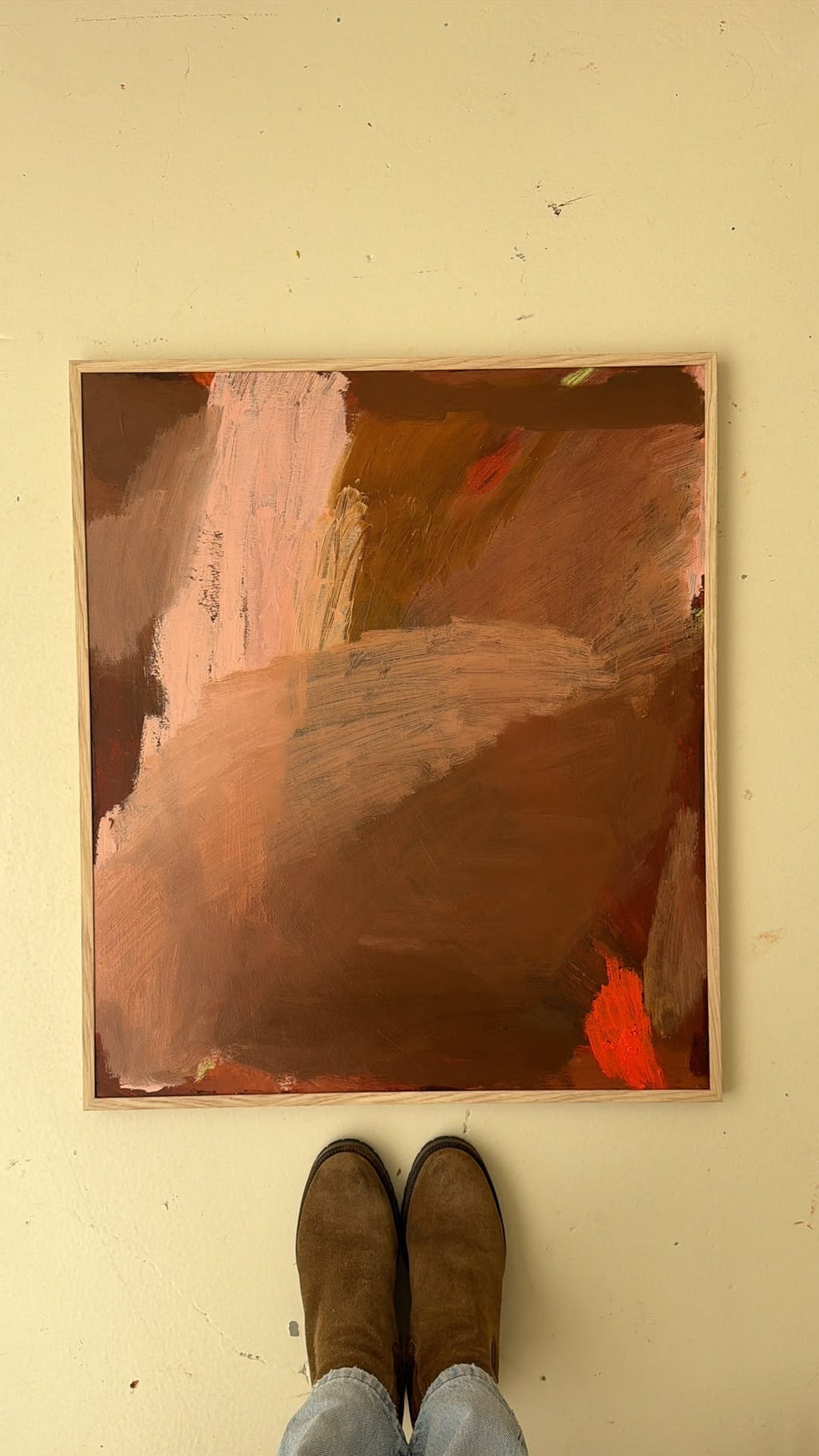 An abstract painting with earthy brown, terracotta, and pink brushstrokes, displayed on a beige wall with a person's feet in brown boots and light blue jeans visible below it.