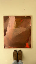 An abstract painting with earthy brown, terracotta, and pink brushstrokes, displayed on a beige wall with a person's feet in brown boots and light blue jeans visible below it.