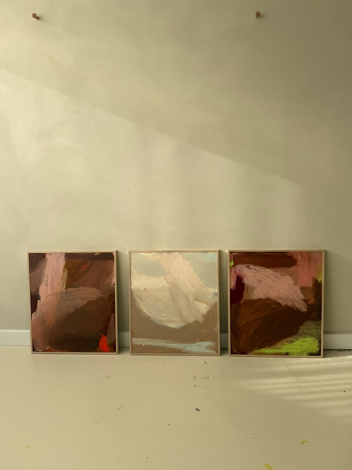 Three framed abstract paintings leaning against a beige wall on a light floor, with warm natural light casting shadows across the wall.