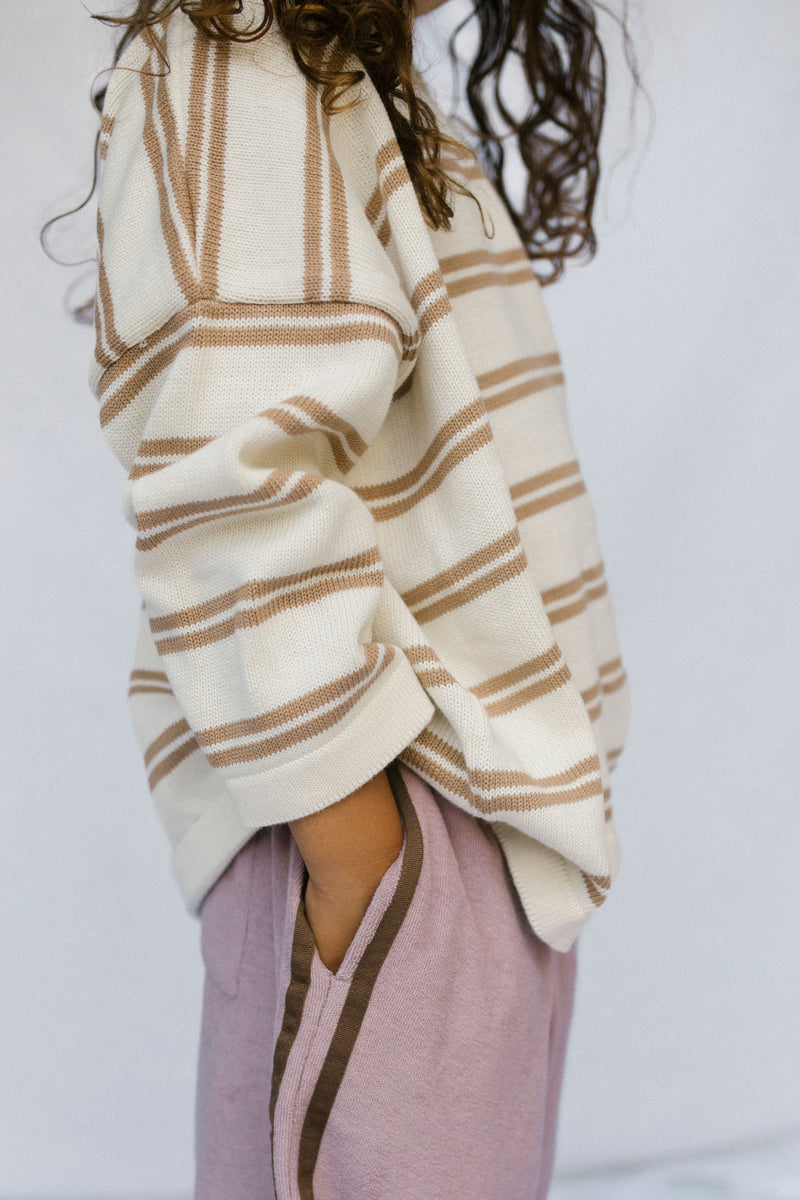 A close-up side view of a person wearing an oversized cream knit sweater with tan horizontal stripes and dusty pink pants with brown side stripes, against a plain light background.