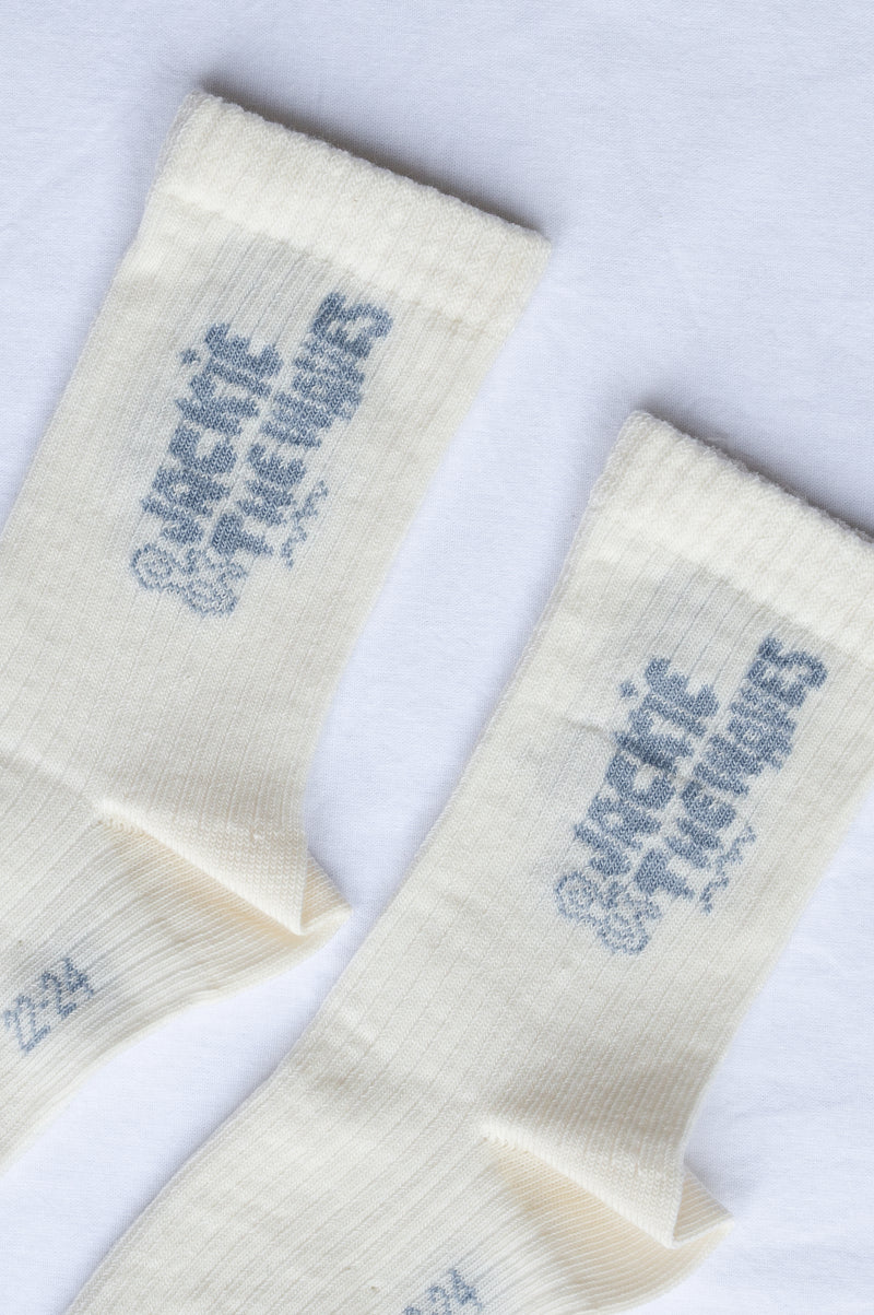A pair of cream-colored ribbed crew socks with a blue graphic print reading 'Jackie DeLuuso's' on the upper cuff, laid flat on a white background.