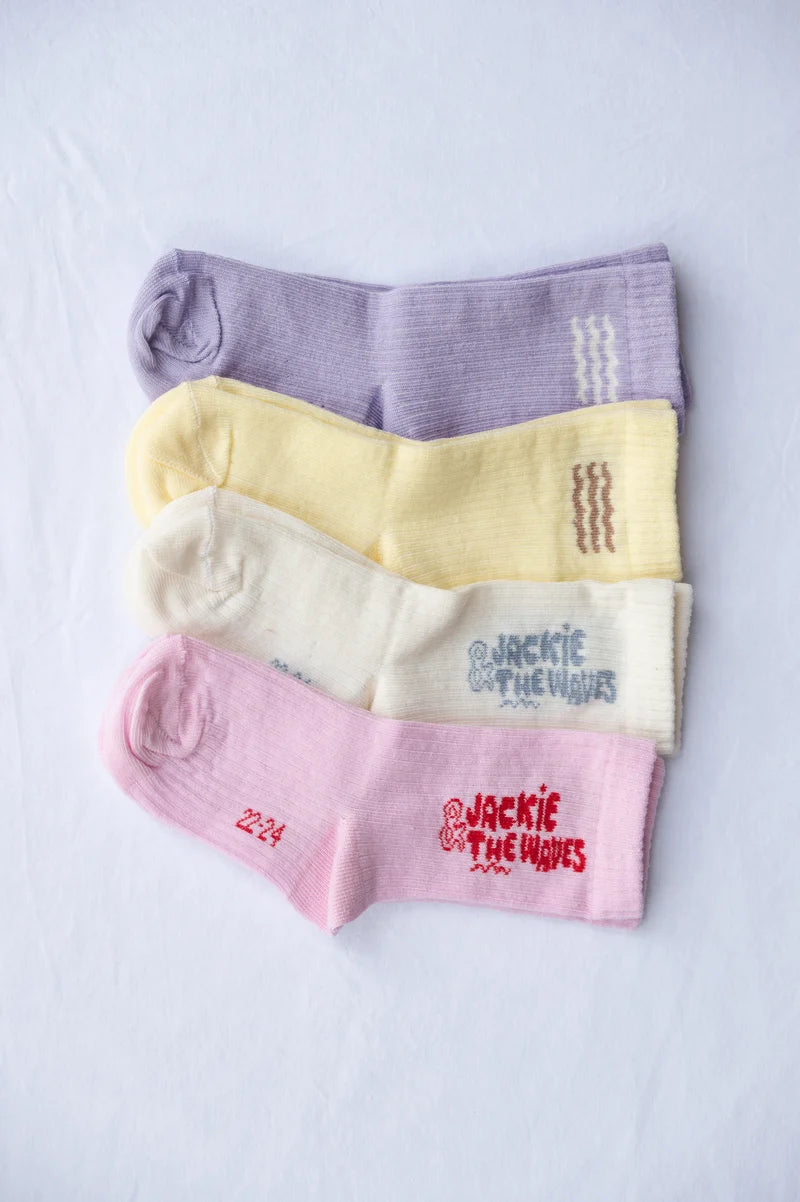 Four pairs of ribbed ankle socks stacked diagonally on a white fabric background. The socks are lavender purple, pale yellow, cream, and light pink, each featuring the 'Jackie Defferre' logo in red on the toe area.