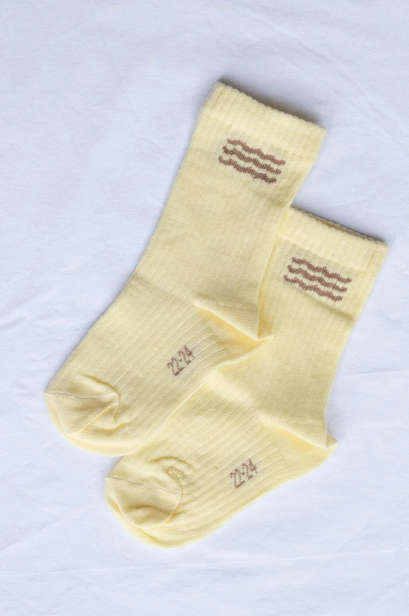 A pair of pale yellow ribbed crew socks with brown wavy stripe details near the cuffs and the number '224' printed on the toes, laid flat on a white fabric background.