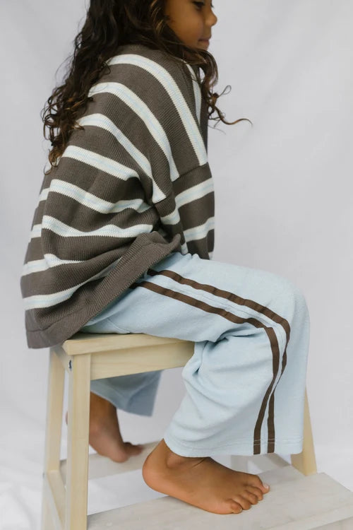 A child seated on a small wooden stool, wearing an oversized brown and white striped knit top and light blue knit pants with dark brown side stripes, against a plain light background.