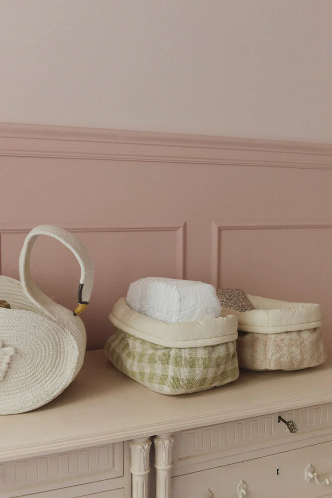 Decorative items including a woven basket, a round container with a checkered pattern, and a teapot on a wooden surface against a pink wall.