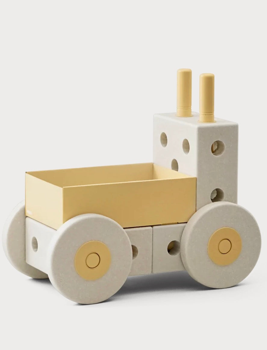 A minimalist wooden toy truck with a cream-colored body, a pale yellow cargo bed, and matching pale yellow details on the wheels and smokestacks, photographed on a light gray background.