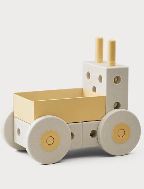 A minimalist wooden toy truck with a cream-colored body, a pale yellow cargo bed, and matching pale yellow details on the wheels and smokestacks, photographed on a light gray background.