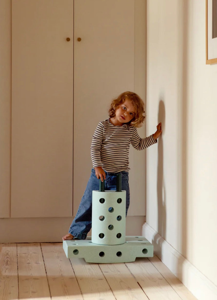 A young child with curly hair stands on a mint green wooden toy platform in a sunlit room with cream-colored walls and wooden floorboards.