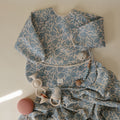 A baby sleep sack with a blue and white floral leaf pattern, styled flat-lay with baby teething toys on a warm cream background.