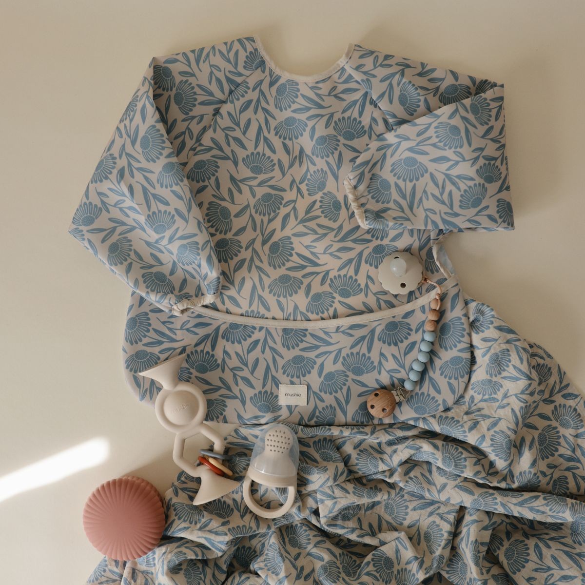 A baby sleep sack with a blue and white floral leaf pattern, styled flat-lay with baby teething toys on a warm cream background.