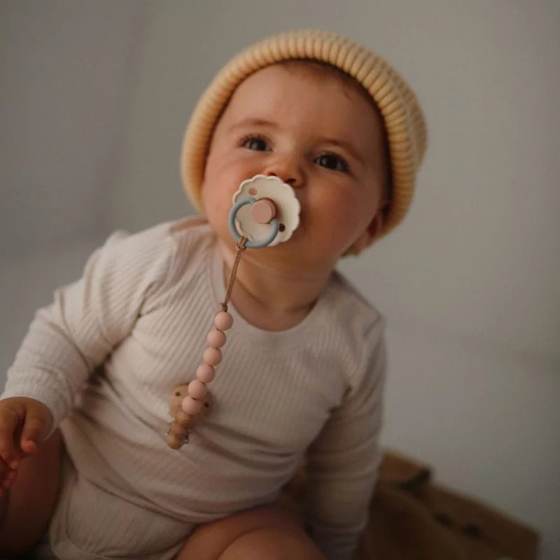 A baby wearing a cream ribbed long-sleeve bodysuit and a beige knit beanie hat, holding a pacifier with a wooden bead chain.