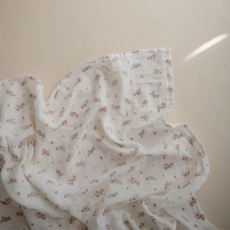 A white muslin baby blanket with a small pink and green floral print, draped in soft folds against a warm beige background.