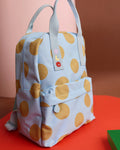 A light blue backpack with large mustard yellow polka dots, featuring dual top handles, a front zip pocket, and a small colorful embroidered patch on the strap, set against a warm pink and orange background.