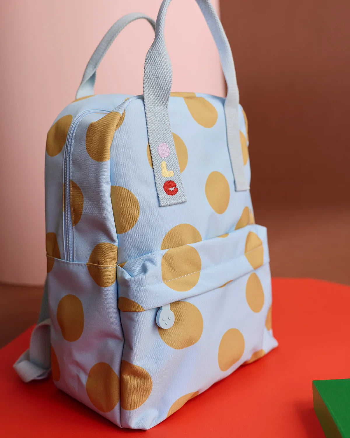 A light blue backpack with large mustard yellow polka dots, featuring dual top handles, a front zip pocket, and a small colorful embroidered patch on the strap, set against a warm pink and orange background.