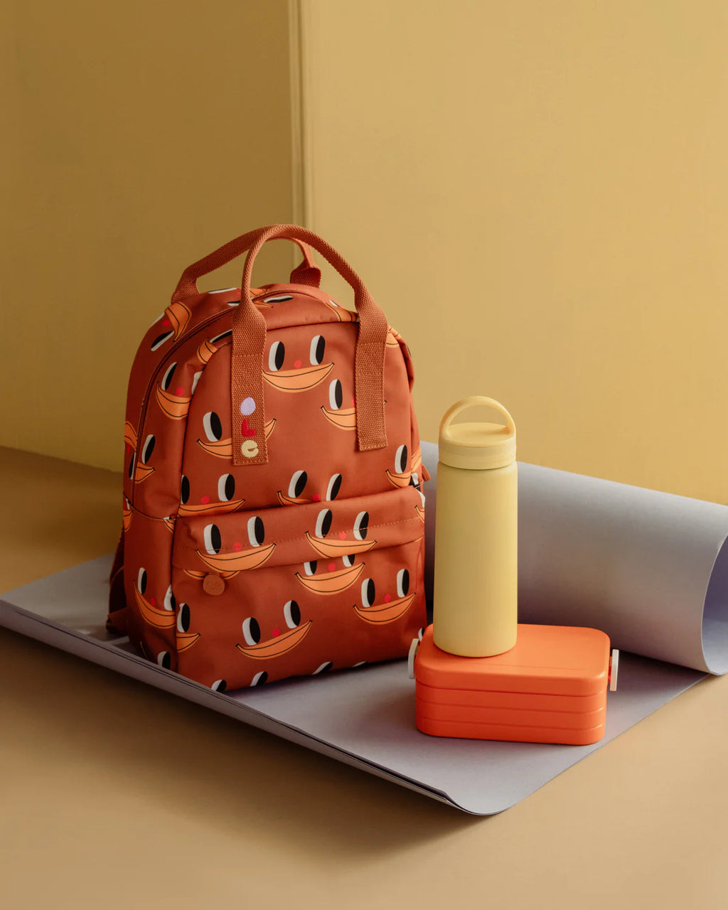 An orange-brown backpack with a repeating banana face pattern, styled on a gray mat next to a pale yellow water bottle and an orange lunch box against a warm beige background.