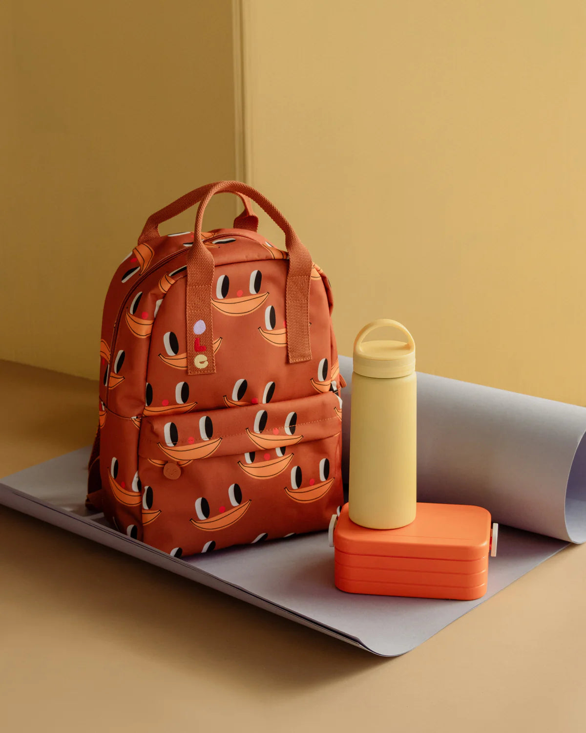 An orange-brown backpack with a repeating banana face pattern, styled on a gray mat next to a pale yellow water bottle and an orange lunch box against a warm beige background.