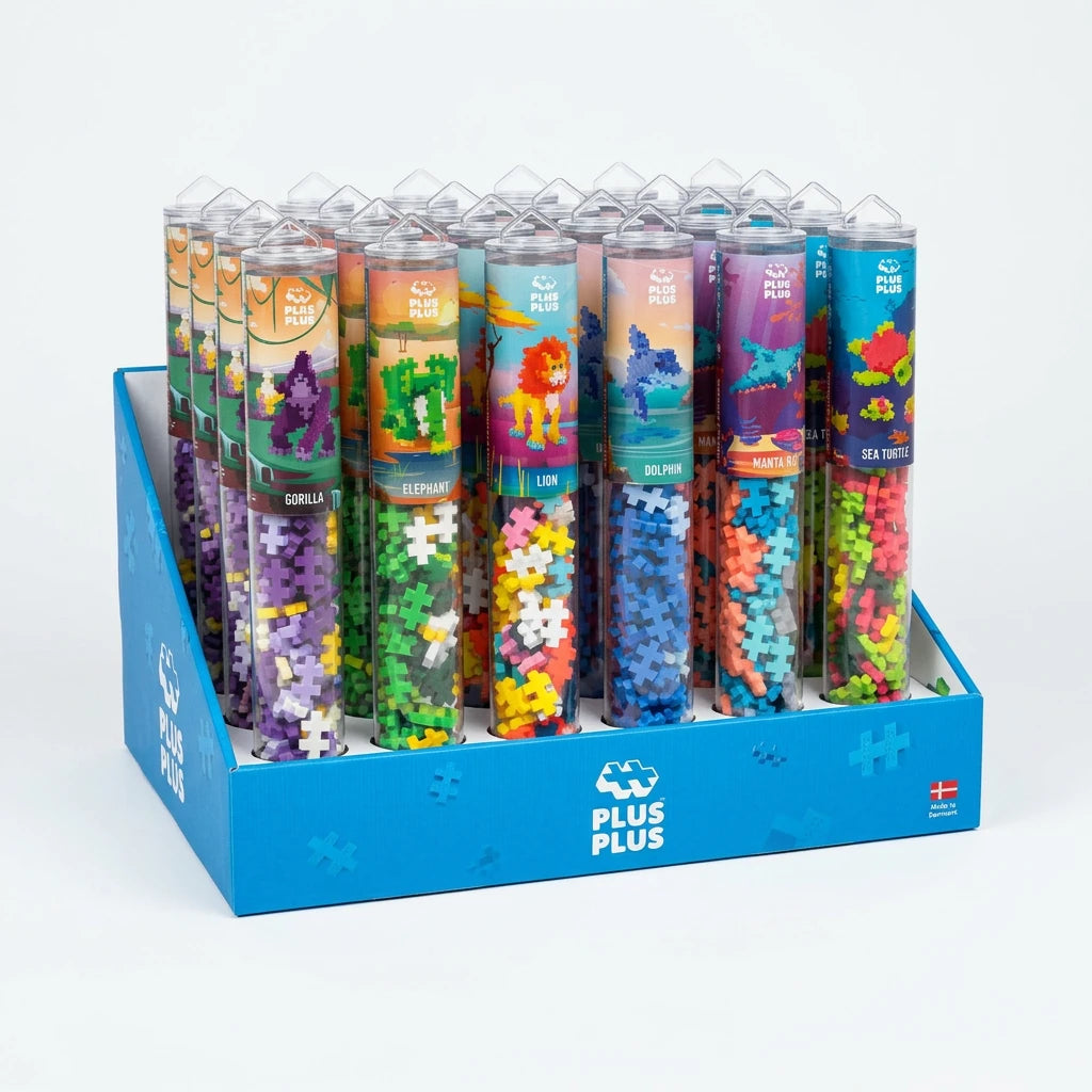A blue PLUS PLUS display box holding eight clear cylindrical tubes filled with colorful jigsaw puzzle pieces. Each tube has a different illustrated label, and the display box is branded with the PLUS PLUS logo.