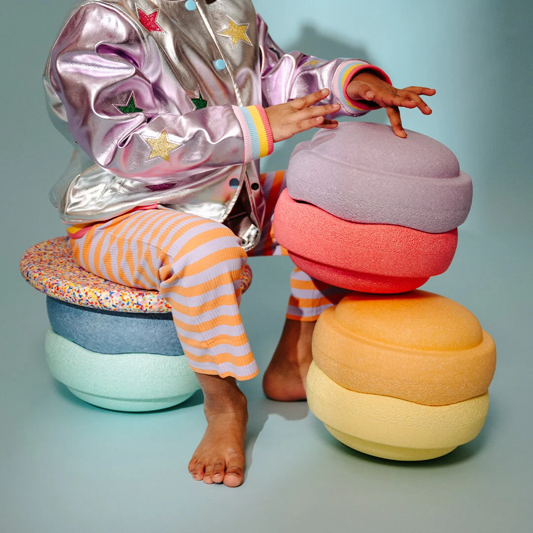 A child wearing a shiny lavender jacket and orange-and-white striped pants sits barefoot on pastel macaron-shaped stools, touching a matching macaron-shaped bench beside them against a pale blue studio background.