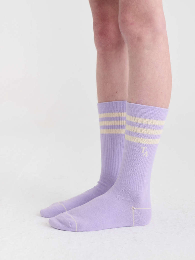 A pair of light purple crew socks with three horizontal cream stripes near the cuff and a small cream logo, shown on a person's feet against a plain light background.