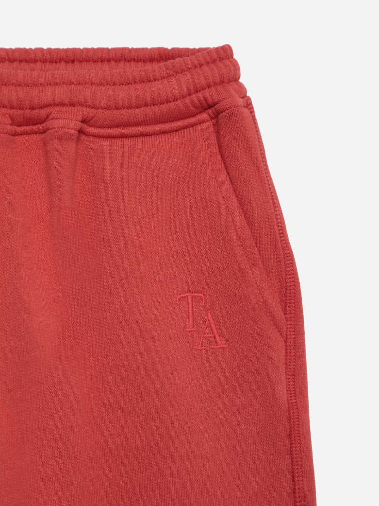 Close-up of a pair of red sweatpants showing the elastic waistband and a tonal embroidered 'TA' logo on the left leg, set against a light gray background.