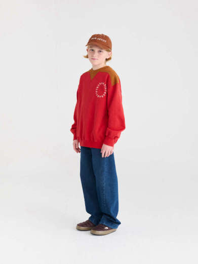 A child model wearing a red crewneck sweatshirt with brown shoulder panels and a white circular graphic on the chest, paired with blue jeans, brown shoes, and a brown cap, standing against a plain white studio background.