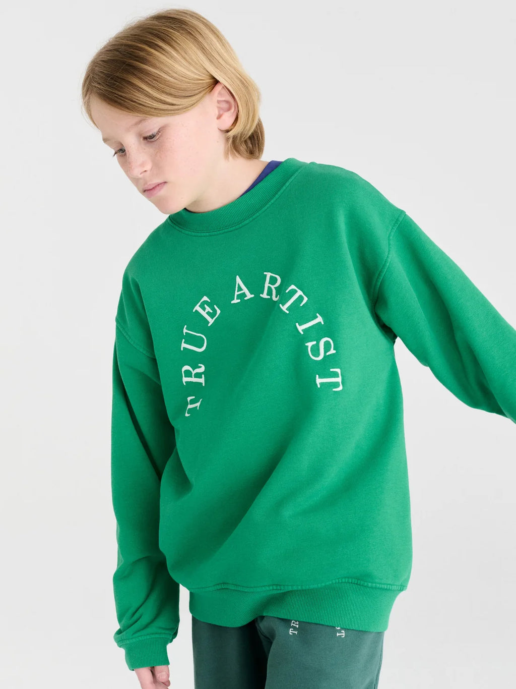 A child wearing a bright green crewneck sweatshirt with 'TRUE ARTIST' printed in white arched text across the chest, photographed against a light gray studio background.