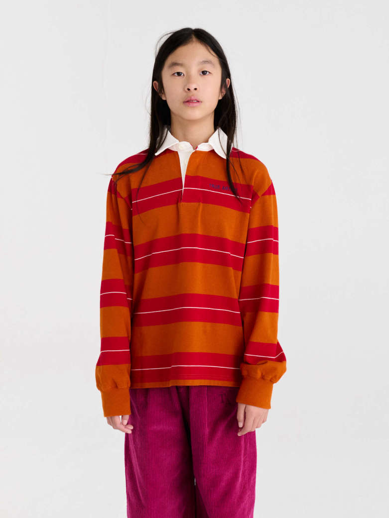 A young girl stands against a light gray studio background wearing a long-sleeve rugby-style top with horizontal orange, red, and white stripes over a white collared shirt, paired with magenta pants.